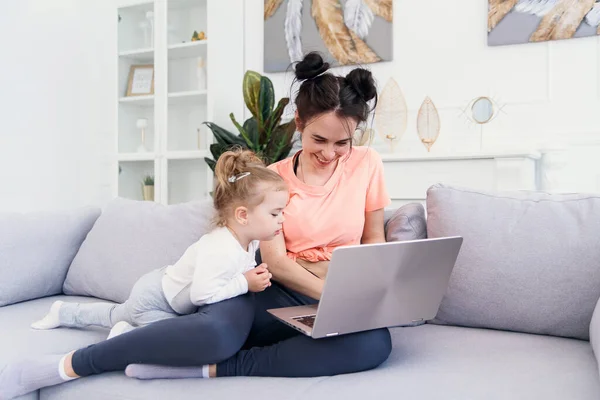 Jovem mãe bonita e sua pequena filha feliz estão usando um laptop e sorrindo enquanto estão sentados no sofá em casa — Fotografia de Stock