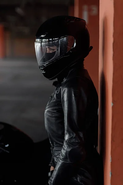 Elegante giovane motociclista donna in abito protettivo nero e casco integrale vicino alla sua moto nel parcheggio sotterraneo. — Foto Stock