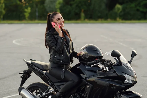 Bonito motociclista feminino fala por smartphone sentado na motocicleta esporte elegante no estacionamento ao ar livre. — Fotografia de Stock