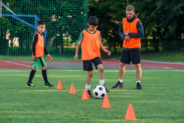 Přetrvávající dospívající chlapec ve sportovním oblečení vlaky fotbal na fotbalovém hřišti a učí se kroužit míč mezi tréninkovými kužely. — Stock fotografie