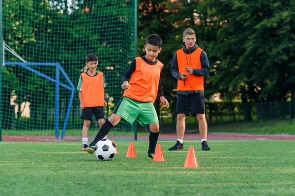 Přetrvávající dospívající chlapec ve sportovním oblečení vlaky fotbal na fotbalovém hřišti a učí se kroužit míč mezi tréninkovými kužely. — Stock fotografie