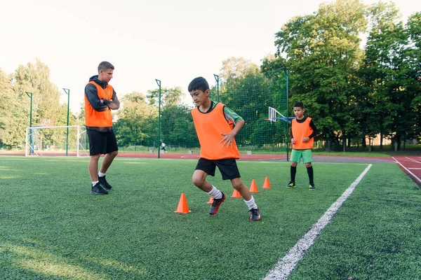 Motivovaný sportovní 13-s chlapci v oranžových vestách běží mezi plastovými kužely během fotbalového tréninku na stadionu. — Stock fotografie