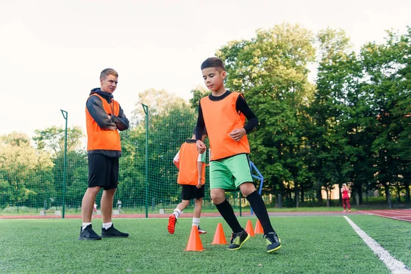 Мотивированные спортивные 13-летние мальчики в оранжевых жилетах, бегающие среди пластиковых конусов во время футбольных тренировок на стадионе. — стоковое фото