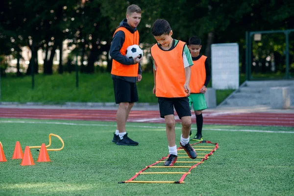 Dva školáci během letního fotbalového tábora cvičí žebříky na trávníku. Intenzivní fotbalový trénink s trenérem. — Stock fotografie