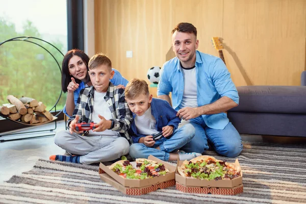 Pai feliz, mãe e dois filhos sentados no chão, jogando videogames com gamepads e comendo pizza saborosa. — Fotografia de Stock