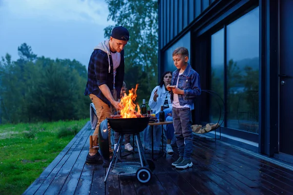 Счастливые отец и сын готовят барбекю на семейном отдыхе на террасе своего современного дома вечером. — стоковое фото