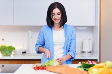 Genç bir kadın ahşap bir tahtanın üzerinde bıçakla lahana keser ve ev mutfağında sebzelerden salata hazırlar..