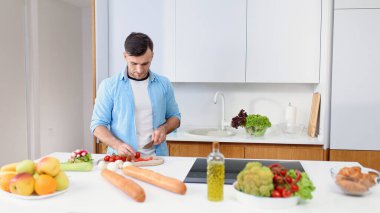 Yakışıklı adam sıcak mutfakta taze sebzelerden salata hazırlamak için ahşap bir tahtada domates kesiyor. Sağlıklı yemek..