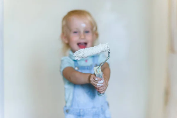 Funny toddler girl holds a white paint roller and smiling in a new house. Selective focus. — Stock Photo, Image