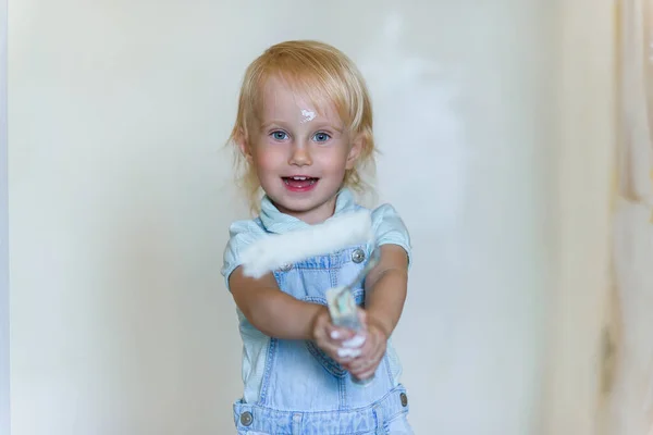 Αστείο κοριτσάκι κρατά ένα λευκό ρολό χρώμα και χαμογελά σε ένα νέο σπίτι. Οικογενειακό διαμέρισμα επισκευής. — Φωτογραφία Αρχείου