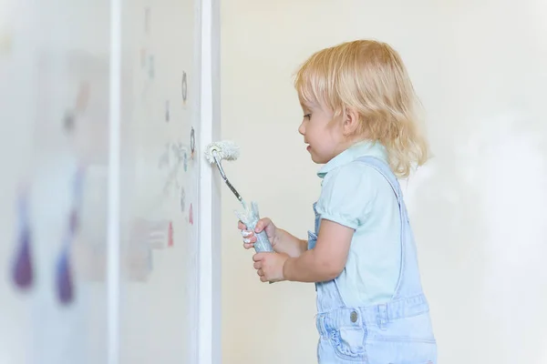 面白い幼児の女の子は新しいアパートで白いペイントローラーで壁をペイントします。家族の修理アパート. — ストック写真