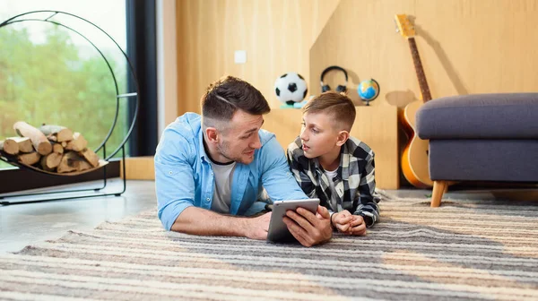 Menino com o pai deitado no chão usando tablet pc no apartamento moderno. Tempo de família feliz juntos. — Fotografia de Stock
