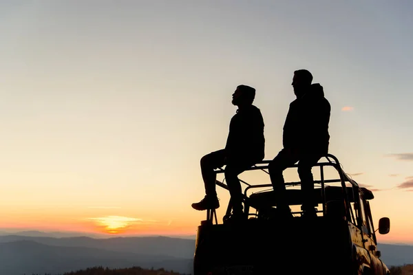 Két felismerhetetlen férfi sziluettje ül egy SUV tetőn egy hegy tetején naplementekor. Extrém utazási koncepció. — Stock Fotó