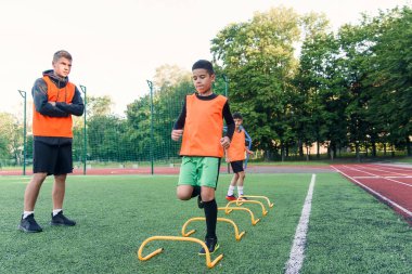 Motive olmuş genç futbolcular koşma egzersizleri yapıyor ve yapay futbol sahasına raflar koyan engelleri aşıyorlar..