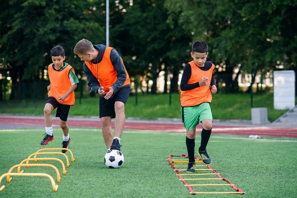 Entrenador de fútbol seguro observa a sus alumnos haciendo ejercicios de carrera con la superación de obstáculos con bastidores en el campo de fútbol — Foto de Stock