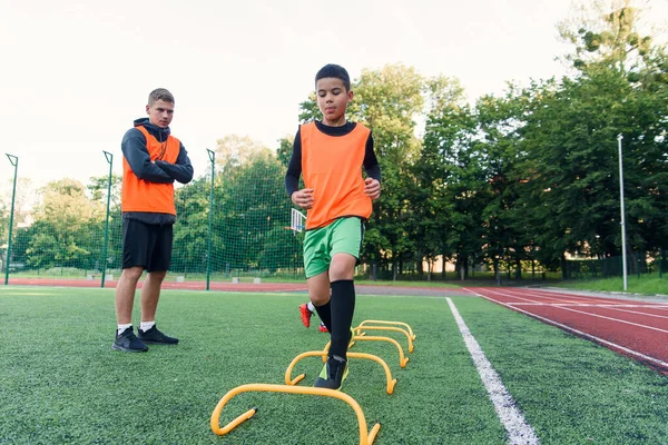 Giocatori di calcio adolescenti motivati eseguono esercizi di corsa con superare gli ostacoli applicando rack sul campo di calcio artificiale. — Foto Stock