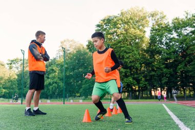 Spor yaparken suni sahada duran turuncu koniler arasında koşan konsantre küçükler futbol oyuncusu.