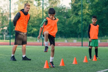 Spor yaparken suni sahada duran turuncu koniler arasında koşan konsantre küçükler futbol oyuncusu.