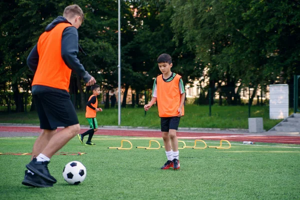 Joueur adolescent habile en uniforme de football travaillant sur le ballon de coups de pied avec son entraîneur expérimenté sur le terrain de sport — Photo