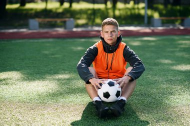 Turuncu yelekli profesyonel futbol koçu suni futbol sahasında otururken elinde bir top tutuyor..