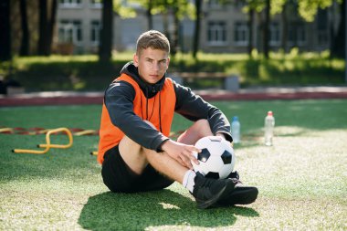 Turuncu yelekli profesyonel futbol koçu suni futbol sahasında otururken elinde bir top tutuyor..