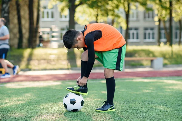 Primer plano joven jugador de fútbol que pone su pierna en la pelota y atar cordones en el estadio de fútbol durante el entrenamiento. —  Fotos de Stock