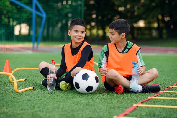 Dois meninos adolescentes felizes em uniformes de futebol tendo um descanso no campo de esporte e desfrutando de água doce no intervalo. — Fotografia de Stock