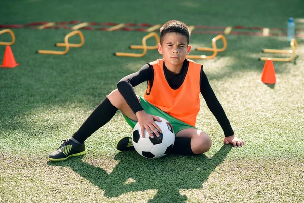 Menino hispânico bonito em uniforme de futebol com bola de futebol após treinamento intensivo no estádio no parque urbano no dia de verão. — Fotografia de Stock