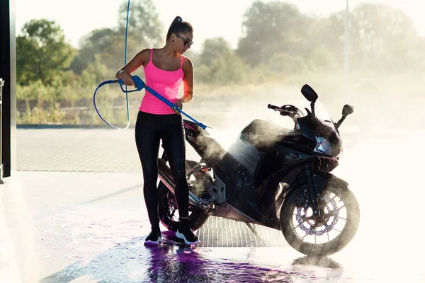 Menina deslumbrante bonita lava uma motocicleta em um auto-serviço com jato de água de alta pressão na parte da manhã ao nascer do sol. — Fotografia de Stock