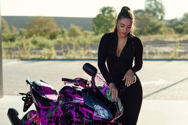 Beautiful girl in tight-fitting seductive suit washes stylish sport motorcycle and wipes it from magenta foam at sunrise. — Stock Photo, Image