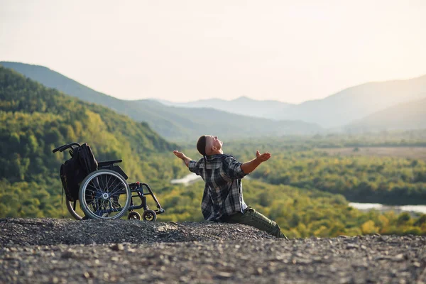 Молодий інвалід сидить на горі біля інвалідного візка і піднімає руки, насолоджуючись свіжим повітрям і красою природи . — стокове фото