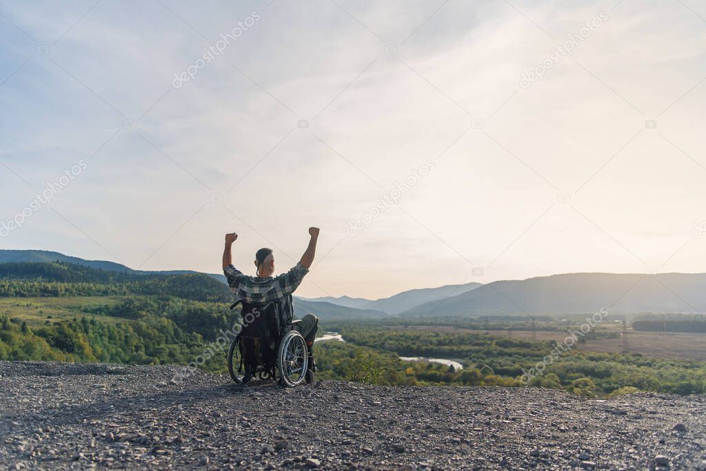 Lonely young man in wheelchair raised hands up rejoicing in his victory on mountain. People with disabilities travel.