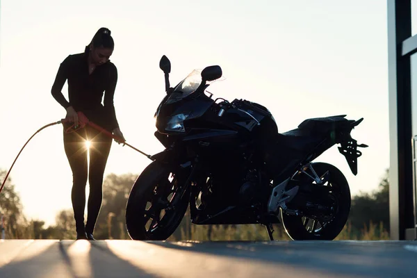 A menina atraente lava uma motocicleta na lavagem de carro de autoatendimento com água de alta pressão. Sol entre pernas. — Fotografia de Stock