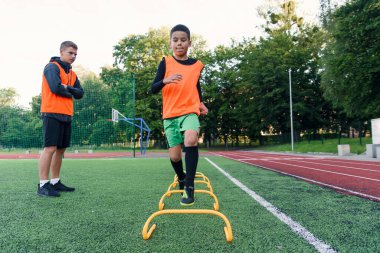 Motive olmuş genç futbolcular koşma egzersizleri yapıyor ve yapay futbol sahasına raflar koyan engelleri aşıyorlar..