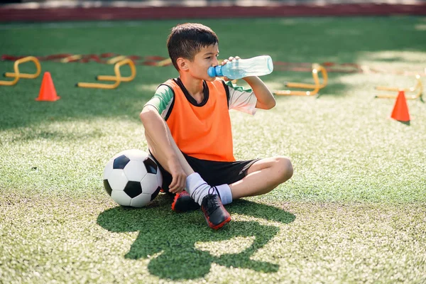 Tired teen boy in soccer uniform drinks with water from plastic bottle after intensive training at stadium in morning. — Stock Photo, Image