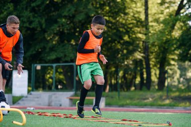 Deneyimli futbol koçu, öğrencisinin futbol stadyumundaki engelleri aşarak koşu egzersizlerini izliyor..