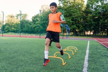 Motive olmuş genç futbolcular koşma egzersizleri yapıyor ve yapay futbol sahasına raflar koyan engelleri aşıyorlar..