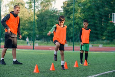Spor yaparken suni sahada duran turuncu koniler arasında koşan konsantre küçükler futbol oyuncusu.