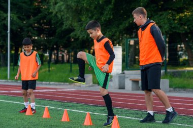 Spor yaparken suni sahada duran turuncu koniler arasında koşan konsantre küçükler futbol oyuncusu.