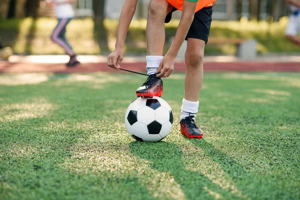 Close up soccer boot of football player which puts his leg on ball and tying shoelace on artificial sports field.