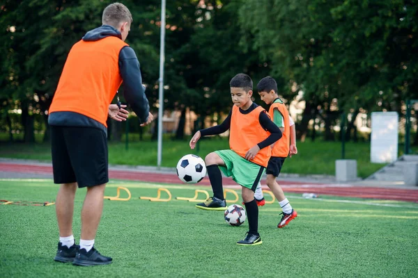 Ragazzo motivato giocatore in uniforme da calcio che lavora fuori la palla calci insieme con il suo allenatore esperto sul campo sportivo — Foto Stock