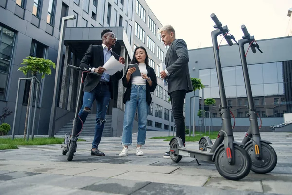El exitoso equipo de negocios multirracial de tres empleados analiza un proyecto conjunto cerca del edificio de oficinas cerca de e-scooters. — Foto de Stock