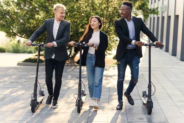 Confiados colegas de oficina multirraciales felices discutiendo proyecto de negocio ir cerca de edificio de oficinas con scooters eléctricos — Foto de Stock
