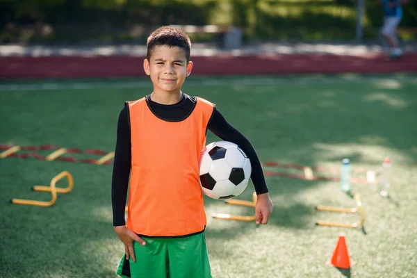 Bonito menino feliz em uniforme de futebol com bola após treinamento produtivo no estádio no parque verde urbano no dia de verão. — Fotografia de Stock