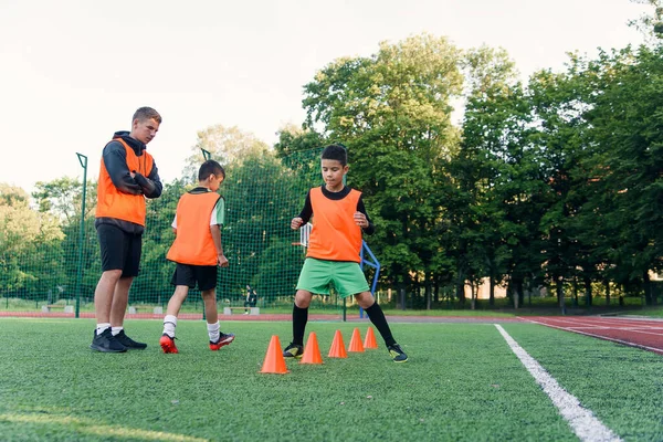 Persistent teen sports boys in orange uniform running among plastic cones during football exercises at the stadium.