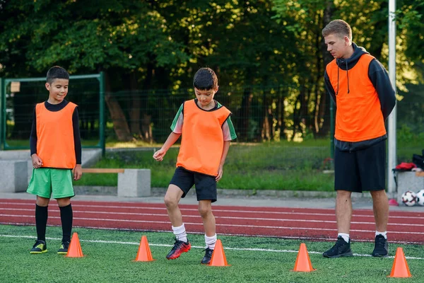 Настойчивые подростки в оранжевой форме бегают среди пластиковых конусов во время футбольных упражнений на стадионе. — стоковое фото