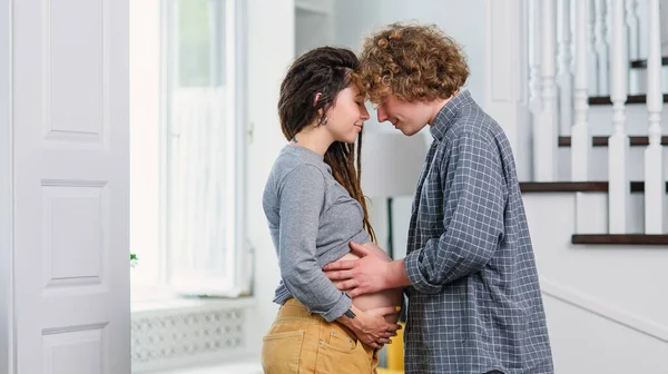 Young hipster pregnant couple standing opposite each other in contemporary apartment and tenderly stroking her belly. — Stock Photo, Image