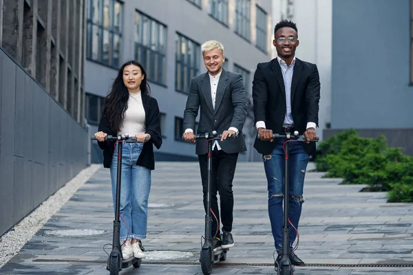 Atractivos colegas de oficina multirraciales alegres montando en scooters eléctricos cerca de un hermoso edificio arquitectónico. — Foto de Stock