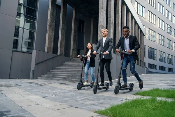 Atractivos colegas de oficina multirraciales alegres montando en scooters eléctricos cerca de un hermoso edificio arquitectónico. — Foto de Stock
