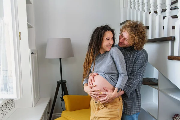 Ευτυχισμένο hipster έγκυος ζευγάρι στέκεται μαζί κοντά σε σκάλες στο σύγχρονο διαμέρισμα και τρυφερά αγγίζοντας κοιλιά. — Φωτογραφία Αρχείου